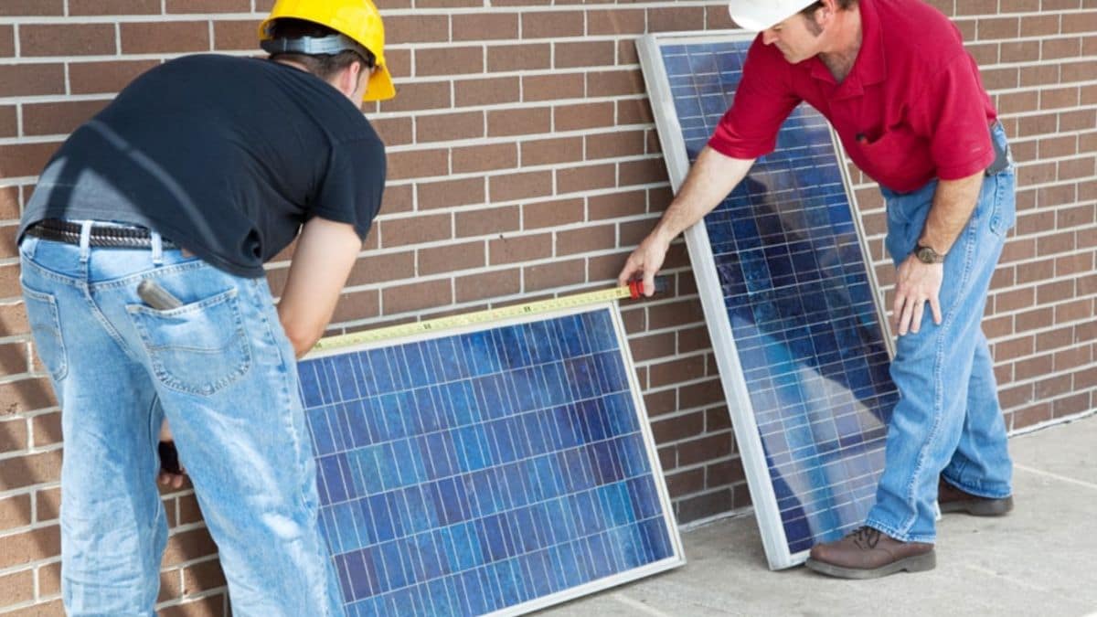 Twee mannen zijn een zonnepaneel aan het opmeten die horizontaal tegen een bakstenen muur aanstaat. Naast die zonnepaneel staat er nog een andere zonnepaneel verticaal tegen de muur. De mannen hebben allebei een helm op