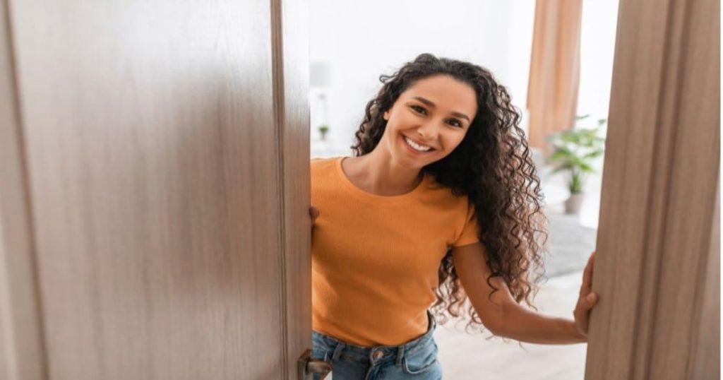Een vrouw met een oranje shirt en krullend haar lacht en opent een Afrormosia houten deur.