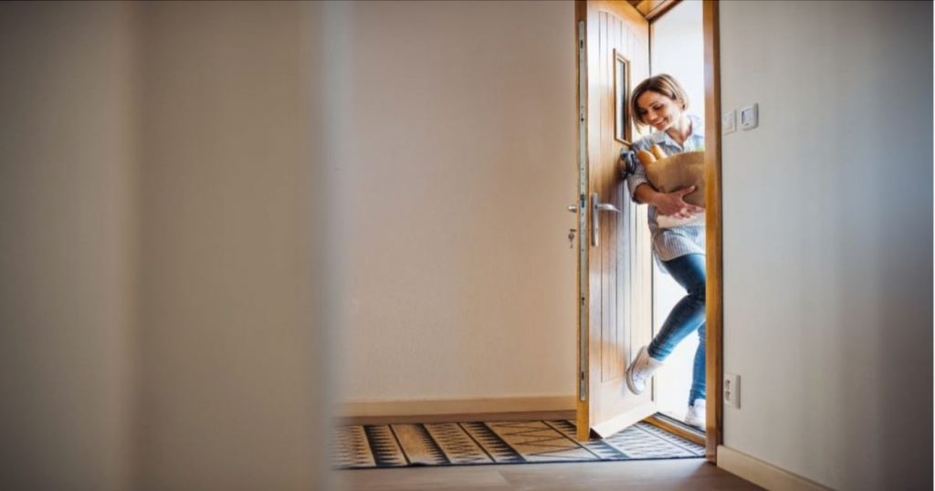 Vrouw duur met haar voet een Afrormosia houten voordeur open terwijl ze een boodschappentas vast heeft. De gang bij de voordeur heeft witte muren en er ligt een mat op de vloer.