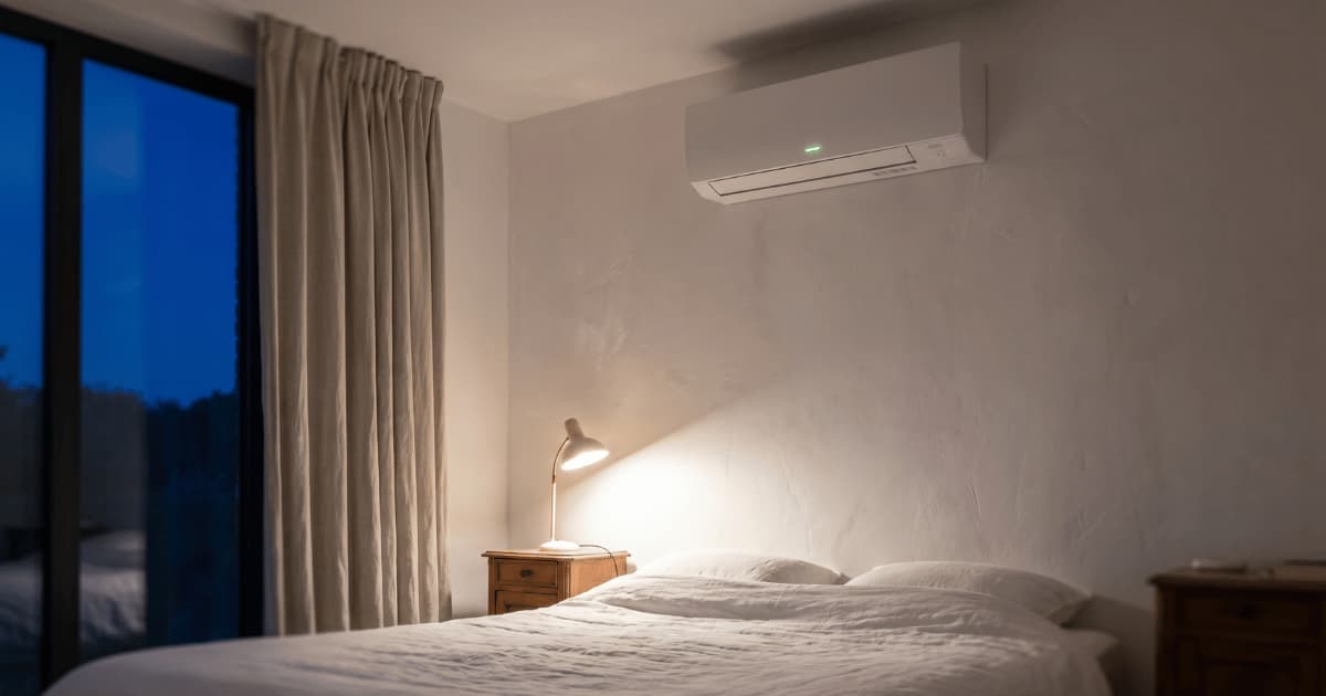 Een witte airco-binnenunit met groen indicatielampje hangt hoog aan de muur boven een bed met wit linnen in een rustige slaapkamer, verlicht door een leeslamp op het nachtkastje en blauw avondlicht door het grote raam.
