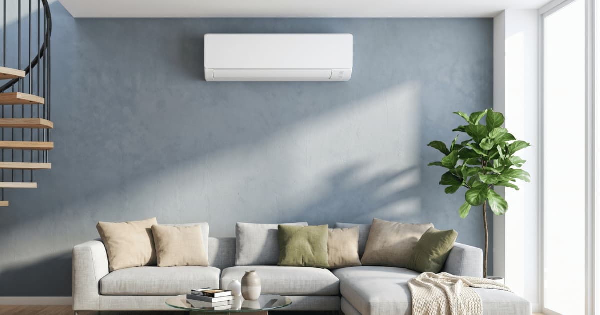 Een witte airco binnenunit hangt hoog aan een blauwgrijze muur boven een lichtgrijze hoekzetel met beige en groene kussens in een moderne woonkamer met een wenteltrap links en een grote plant rechts.