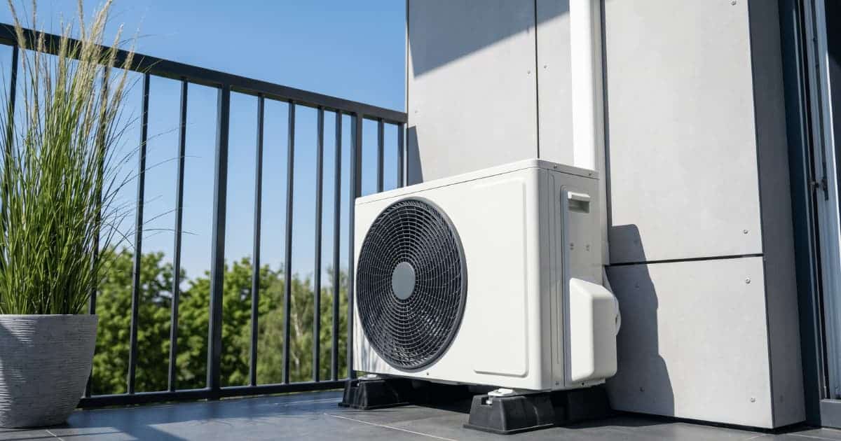 Een witte airco-buitenunit staat op zwarte rubberen trillingsdempers in de hoek van een modern appartementsbalkon met donkere tegels, een metalen balustrade en een siergras in een grijze pot ernaast.