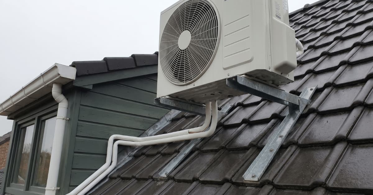 Een witte airco-buitenunit staat op een gegalvaniseerde stalen dakconsole op een hellend dak met donkere betonpannen, terwijl witte geïsoleerde koelleidingen over de pannen naar een groengrijze houten dakkapel lopen.