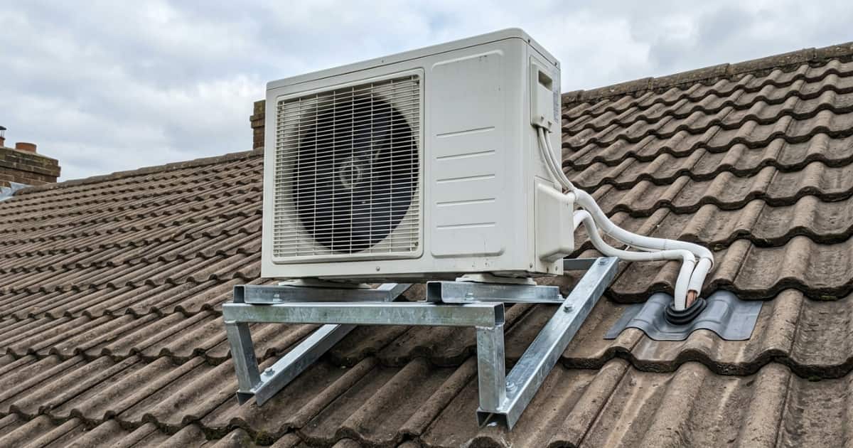 Een witte airco buitenunit staat op een gegalvaniseerd stalen montageframe dat over meerdere rijen bruingrijze keramische dakpannen is bevestigd, met witte geïsoleerde koelleidingen die via een doorvoerpan het dak in verdwijnen.