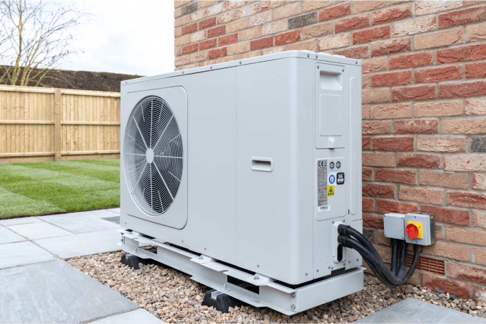 airco buitenunit naast een bruine stenen muur. De buitenunit staat in de tuin van een woning.