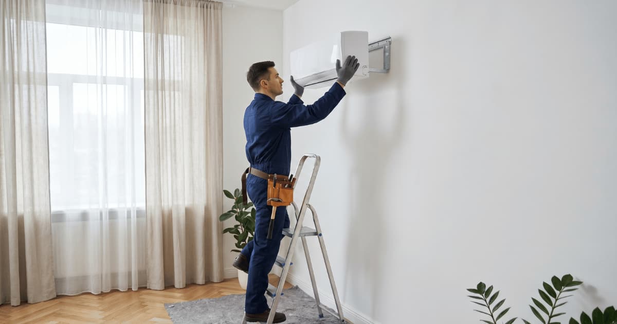 Een installateur in blauwe werkkleding staat op een trapladder en bevestigt een witte airco-binnenunit hoog aan de muur in een lichte kamer met gordijnen op de achtergrond.