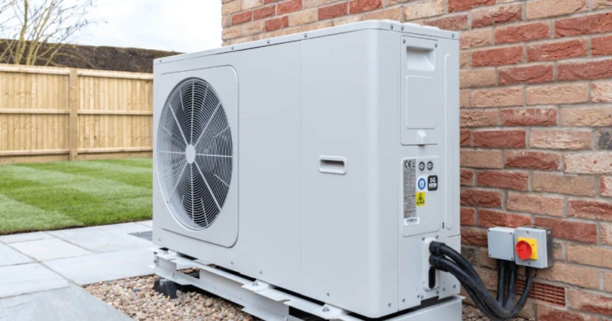 De buitenunit van een airco in de tuin, vlak tegen een muur met bruin en beige bakstenen.