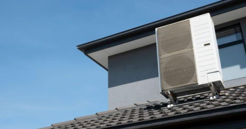 Buitenunit van een airco op een schuin dak net voor een geblindeerd venster en een grijze muur.