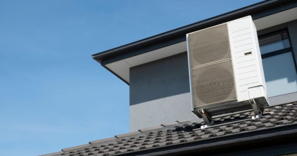 Buitenunit van een airco op een schuin dak net voor een geblindeerd venster en een grijze muur.