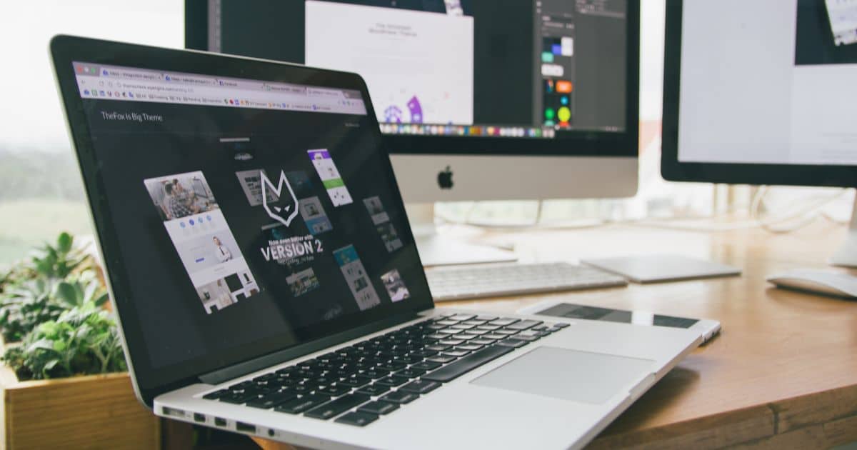 Een geopende laptop voor twee grote monitors. Op het scherm het webdesign en het ontwerp voor de huisstijl van een nieuw bedrijf.