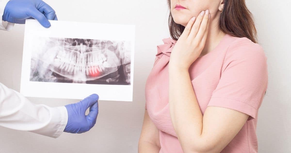 Een dame met bruin haar en een roze t-shirt houdt haar hand tegen haar kaak. De bovenste helft van haar gezicht is niet zichtbaar. Langs haar rechterkant toont een tandarts met witte jas en blauwe handschoenen een röntgenfoto van haar gebit, waarop in het rood een abces te zien is. Enkel de armen en handen van de arts zijn zichtbaar.