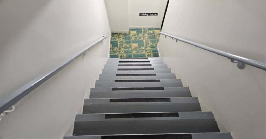 Een rechte, grijze trap met antislip: langs beide kanten is een trapleuning voorzien. Beneden is een groen-gele vloer zichtbaar: tegels of vinyl?