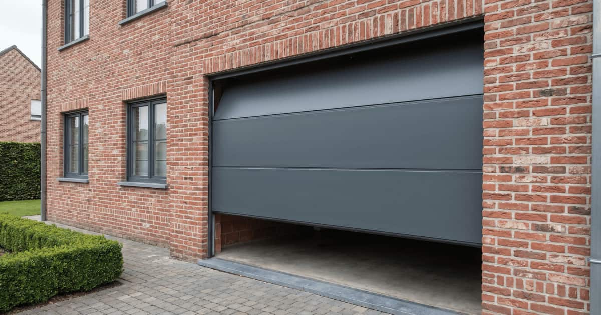 Een antracietgrijze sectionaalpoort schuift naar boven langs de geleiderails aan een Vlaamse bakstenen woning met donkere raamprofielen en een verzorgde klinkeroprit op een bewolkte dag.
