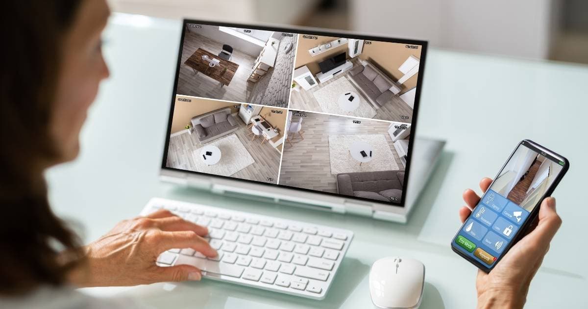 Een dame zit voor een computer met wit toetsenbord en witte muis. Ze heeft haar linkerhand op het toetsenbord. In haar rechterhand houdt ze een smartphone. Op het scherm van de computer houdt ze 4 ruimtes in haar woning in de gaten. Op het scherm van haar smartphone is een vijfde ruimte zichtbaar. De beelden zijn afkomstig van haar beveiligingscamera's.