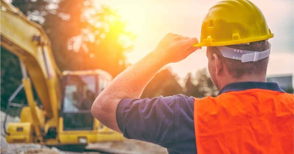 Een gespecialiseerd grondwerken (met gele veiligheidshelm en een blauw hemd onder een oranje fluohesje) kijkt naar een gele rupskraan op een bouwwerf.