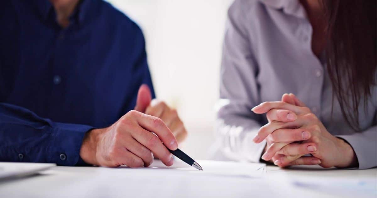 Twee mensen (links een man in blauw hemd, rechts een dame in lichtpaars hemd) zitten naast elkaar aan een witte tafel. Enkel hun bovenlichamen zijn zichtbaar. De heer wijst iets aan op een blad, met een zwarte pen met zilveren top. De dame heeft haar handen in elkaar geslagen.