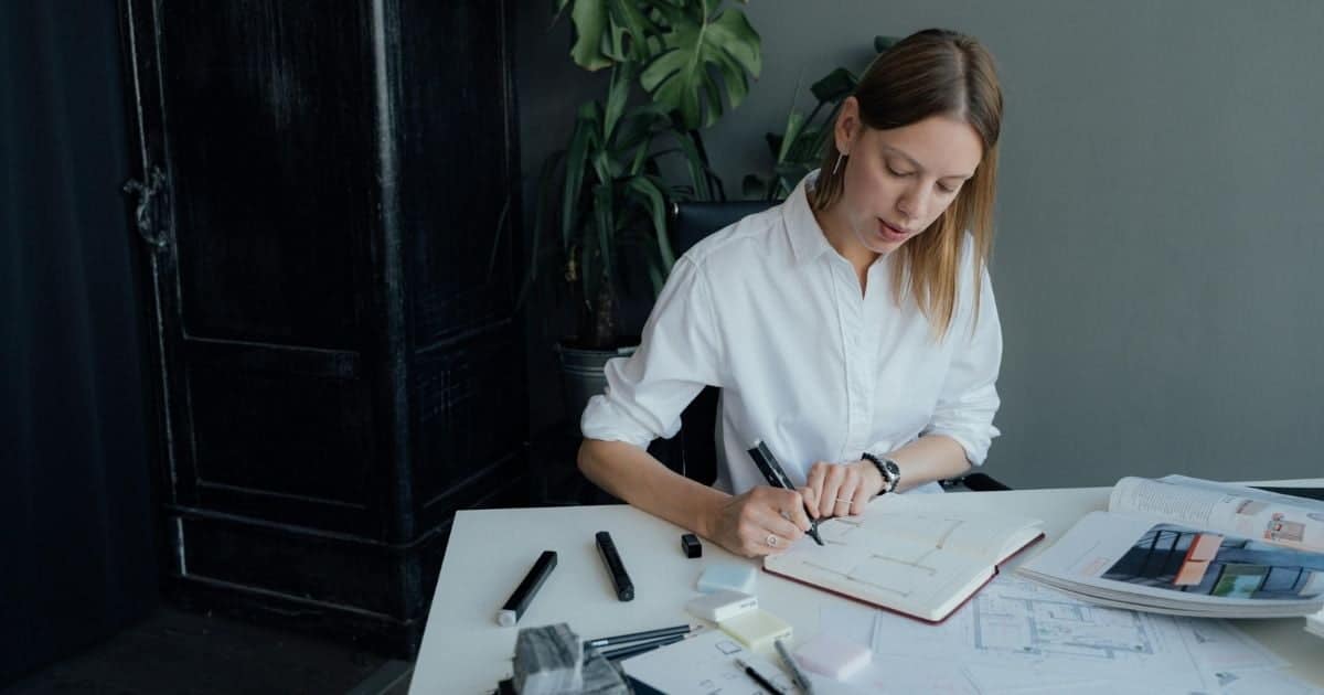 Een architecte werkt in een stijlvol ingerichte kamer met groengrijze wand, donkere, houten antieken kast en monstrera. Ze draagt een wit hemd en noteert iets in een notitieboekje. De architect zit aan een tafel met wit tafelblad, bezaaid met tekeningen, schrijf- en tekengerei, en een boek met afbeeldingen.
