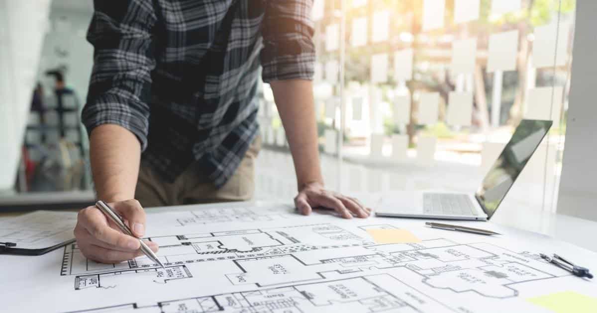 Een architect in grijsblauw geruit hemd buigt zich over een plan of bouwtekening van een woning. In zijn rechterhand heeft hij een pen vast. Rechts van hem, op het plan, staat een grijze laptop.