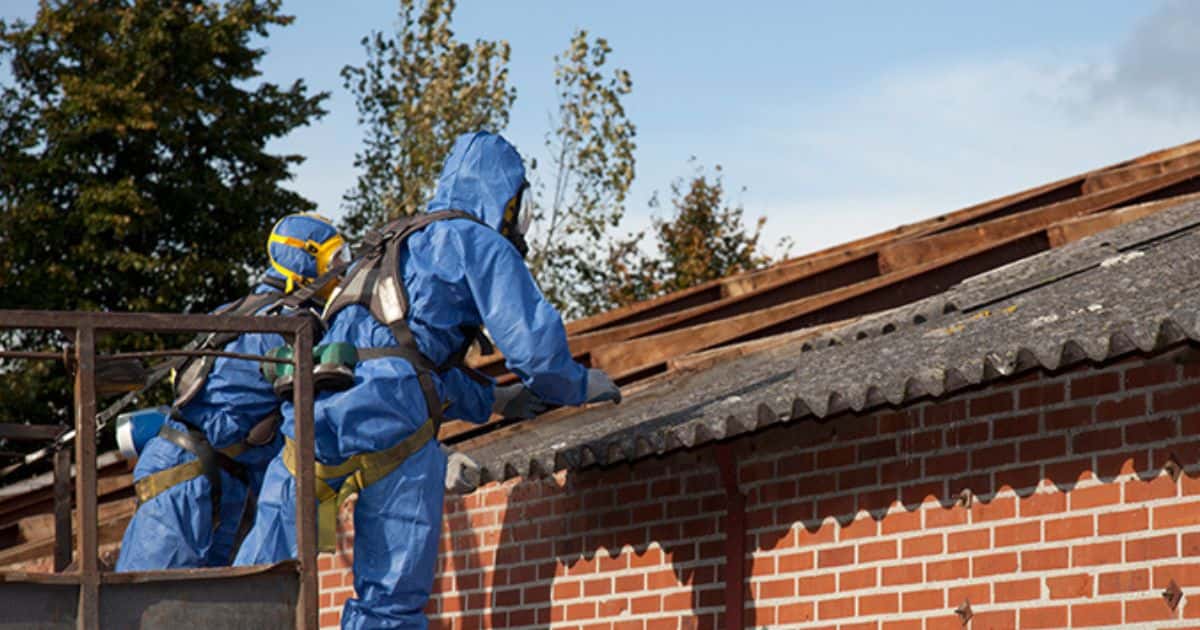 Professionele asbestverwijderaars in blauwe, beschermende kledij en met mondmasker zijn bezig met het verwijderen van asbestplaten op een schuin dak.