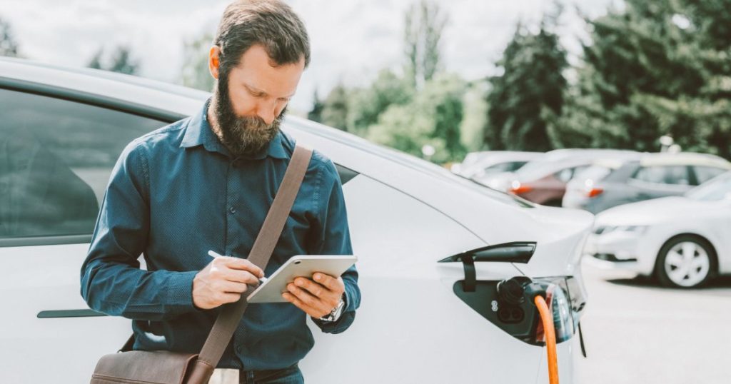 Een man met baard in blauwe overall heeft een tablet vast en een leren tas rond zijn schouder. Hij leunt tegen een witte elektrische auto, die via een oranje laadkabel is aangesloten op een laadpaal.