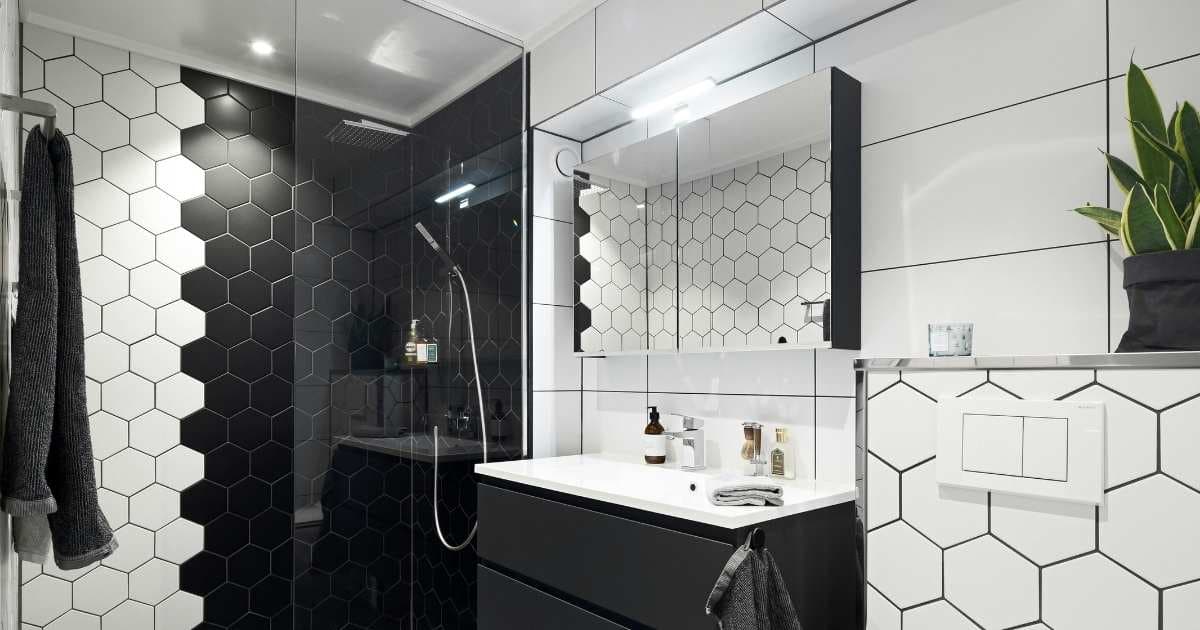 Een moderne badkamer met vijfhoekige tegels. Het leeuwendeel van de badkamer is afgewerkt met witte tegels. In de douche (met glazen wand) is een deel van de wand betegeld met zwarte tegels. In de badkamer staat een wastafel met daarboven een spiegel.