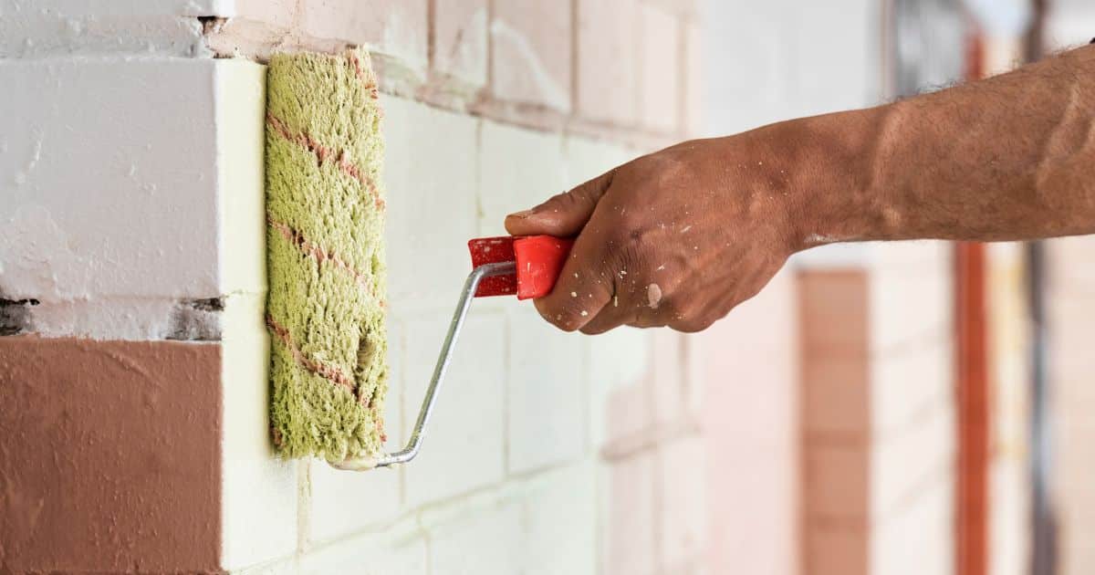 Schilder aan het werk met gelige verfrol met rood handvat op een bakstenen muur.