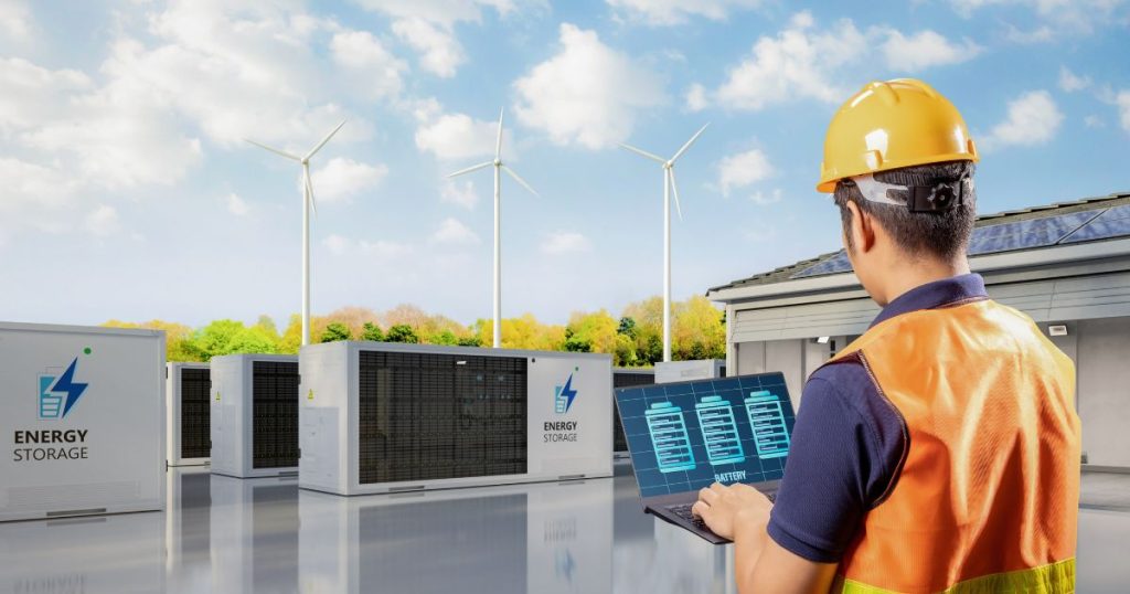 Ingenieur met gele helm en oranje fluohesje controleert een energieopslagsysteem van verschillende batterijen. Op de achtergrond staan drie windmolens.