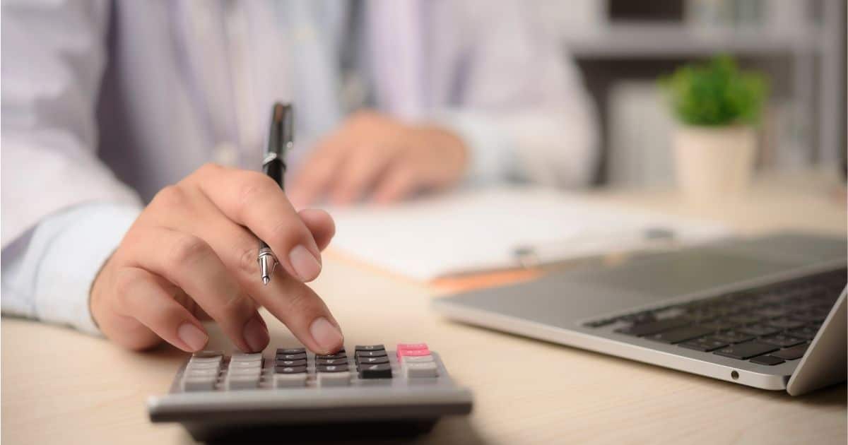 Een hand met middelvinger op een knop van een grijs rekenmachine. Tussen middel- en wijsvinger zit een zwarte pen. Op de achtergrond de borst en andere hand van een man in doktershemd, een laptop, kamerplantje en klembord met papieren.