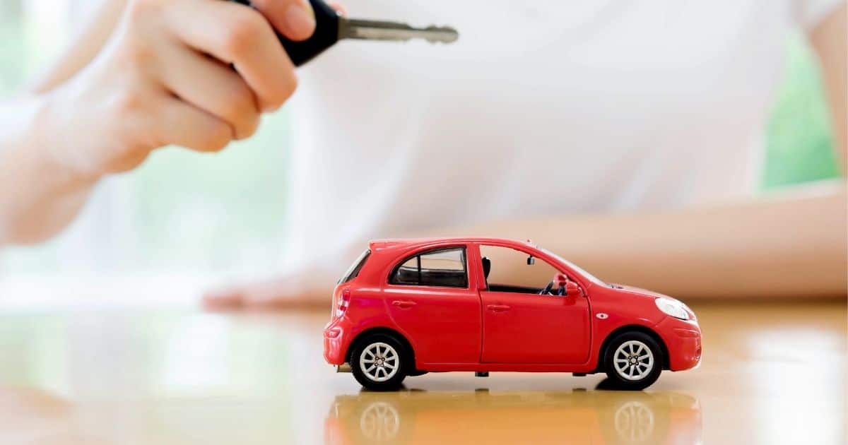 Een rode miniatuurauto op een glanzende houten tafel. Achter de tafel zit een persoon in wit t-shirt. De persoon houdt een autosleutel boven de miniatuurauto.