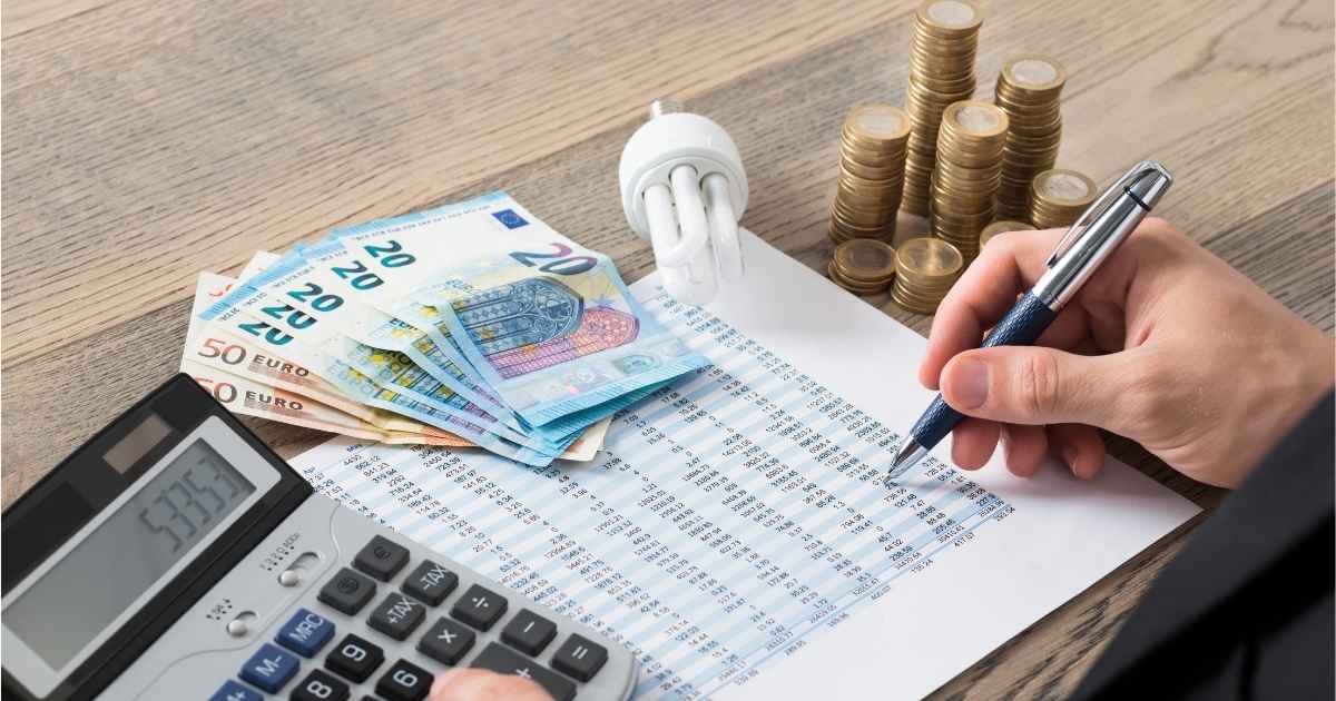 Een woningeigenaar berekent de kosten voor nieuwe isolatie. Hij gebruikt een rekenmachine, pen en tabel. Op de houten tafel liggen ook eurobiljetten, stapels euromunten en een spaarlamp.