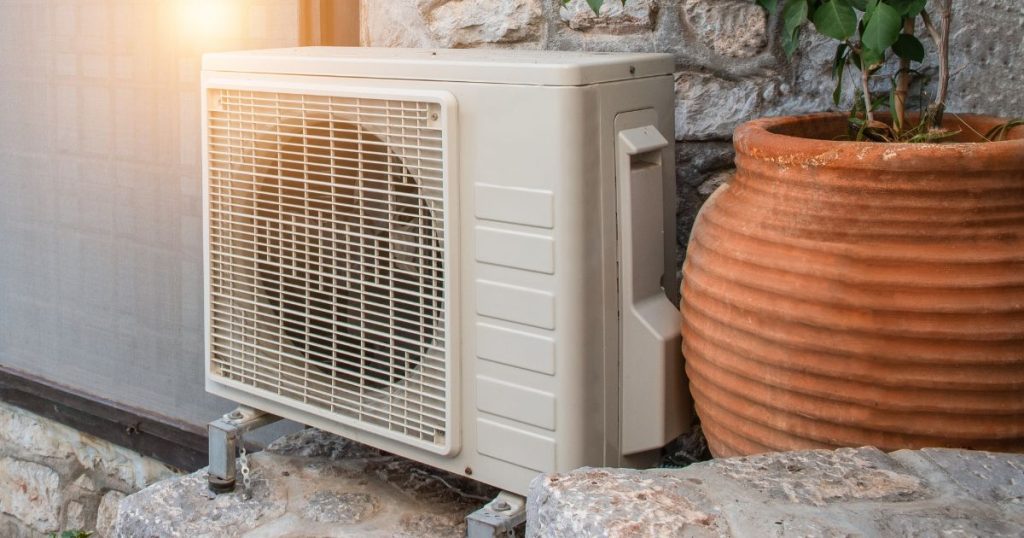 Een warmtepomp tegen de stenen gevel van een woning. Naast de warmtepomp staat een bolle, terracotta bloempot.