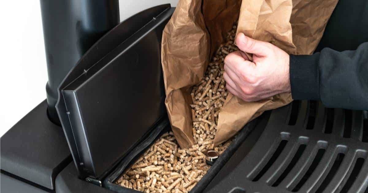De handen van een man gieten pellets uit een papieren zak in een zwarte gietijzeren pelletkachel.