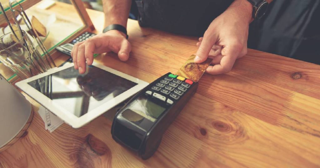 Een man gebruikt een betaalterminal gelinkt aan een iPad kassa om een betaling te verwerken. De witte iPad kassa en zwarte betaalterminal staan op een houten toonbank.