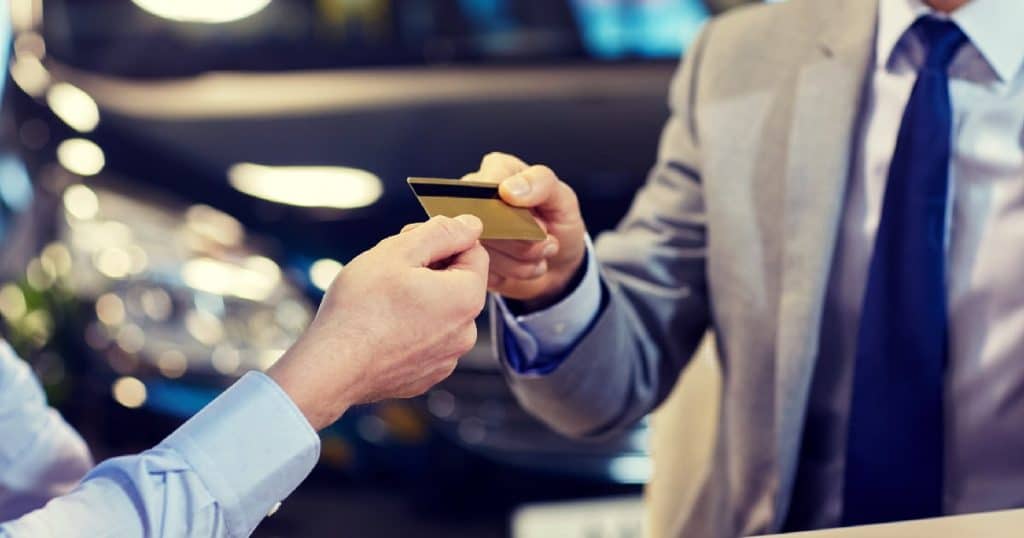 Een man in een grijs pak met een blauwe das overhandigt zijn kredietkaart aan een verkoper in een lichtblauw hemd. Op de achtergrond blinkt een donkerblauwe wagen.