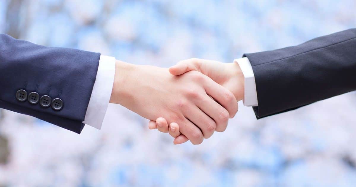 Twee mensen schudden elkaar de hand. Enkel hun armen en handen zijn zichtbaar. De linkse draagt een blauw vest met knopen boven een wit hemd. De rechtse draagt een zwart vest boven een wit hemd.