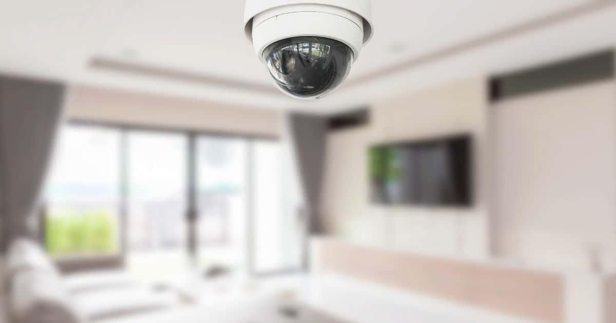 Een dome-camera hangt tegen het plafond van een woonkamer. De woonkamer is vervaagd in beeld: er hangt een televisie aan de muur, er staat een witte zetel, en de ruimte heeft glazen schuifdeuren.