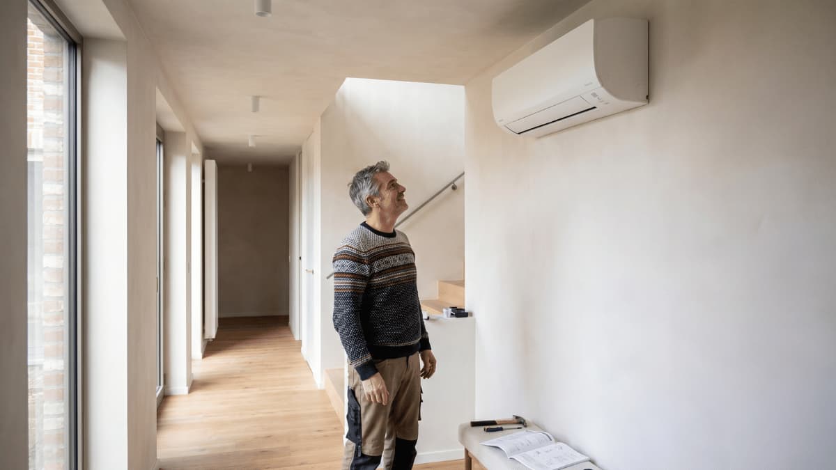 Een man met grijs haar en een gebreide trui staat in de hal van een moderne nieuwbouwwoning en kijkt omhoog naar een witte split-airco binnenunit aan de muur, met een open trapopgang achter hem en smalle raampartijen die baksteendetails tonen.