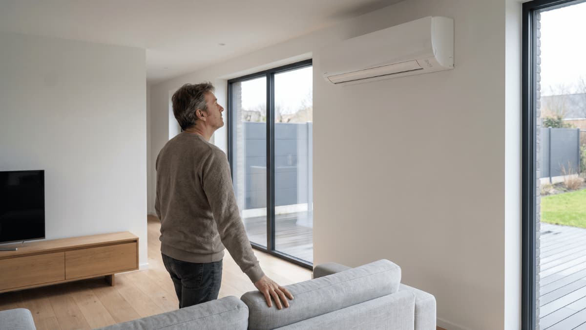 Een man in een beige trui staat naast een lichtgrijze zitbank in een moderne woonkamer en kijkt omhoog naar een witte split-airco binnenunit, met grote schuiframen die uitkijken op een terras met composiet vlonders en een antracietkleurig tuinscherm.