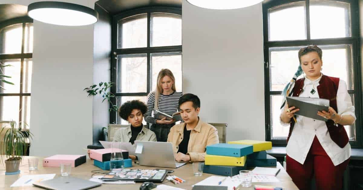 Een groep werknemers, zittend en staand, aan een tafel die bezaaid is met documenten, post-its, geelblauwe dozen, laptops en fardes.