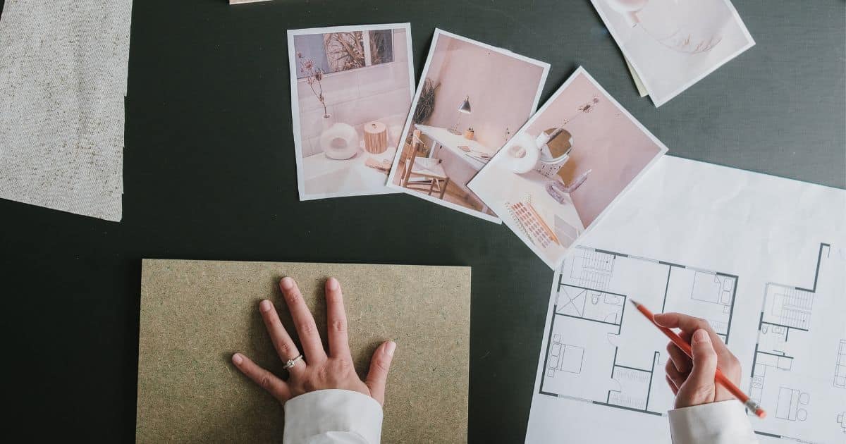 De handen van een interieurarchitect boven een zwarte tafel met een plattegrond, houten textuurstaal, een textielstaal, en drie foto's voor een moodboard.