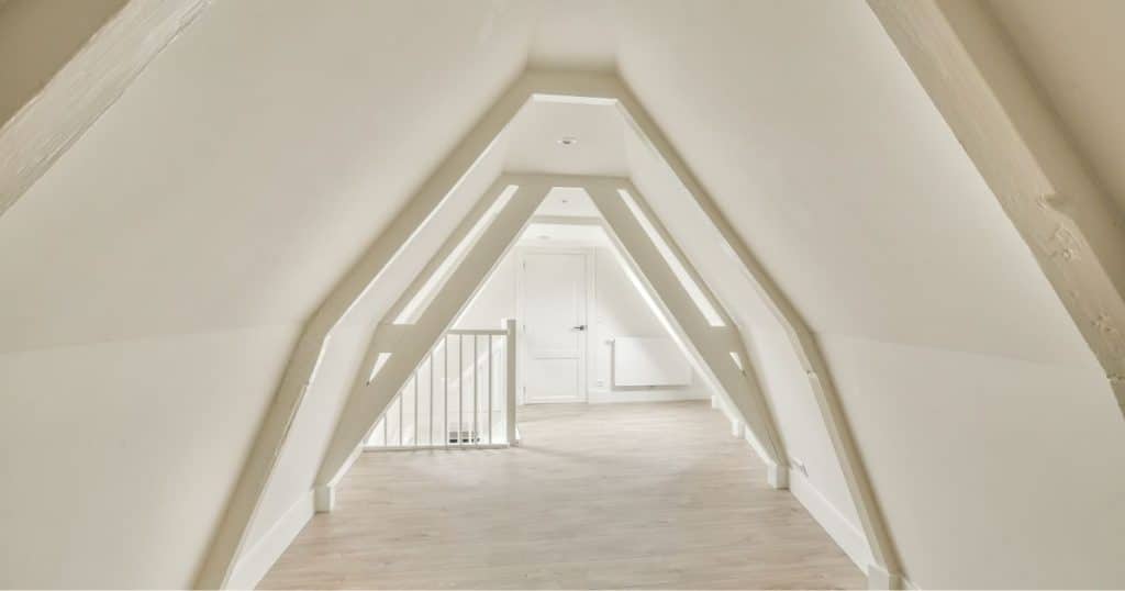 Binnenkant van een zolder onder een smal mansardedak, afgewerkt met witte pleister en een houten vloer.