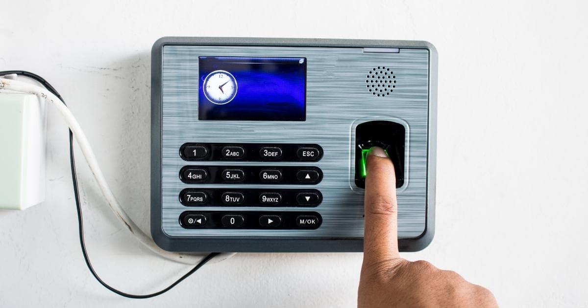 Een vinger op de vingerafdruklezer van een biometrisch tijdsregistratiesysteem. De terminal is grijsgroen, en heeft een klein scherm met een klokje tegen een blauwe achtergrond. Het alfanumerieke toetsenbord is zwart, de cijfers en letters zijn wit.