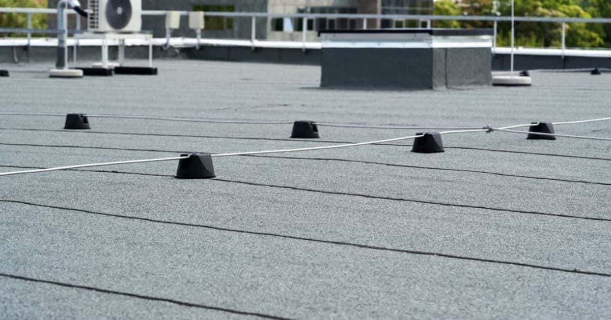Grond van een dak bedekt met bitumen. Op een verhoog staat de buitenunit van een airco (of warmtepomp?). Ook staan er podjes met bekabeling op het bitumen dak. 