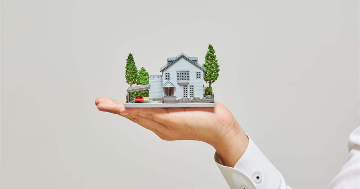 Een hand in witte hemdsmouw presenteert een maquette van een blauwe woning met carport en hoge bomen. Op de achtergrond een grijswitte muur.