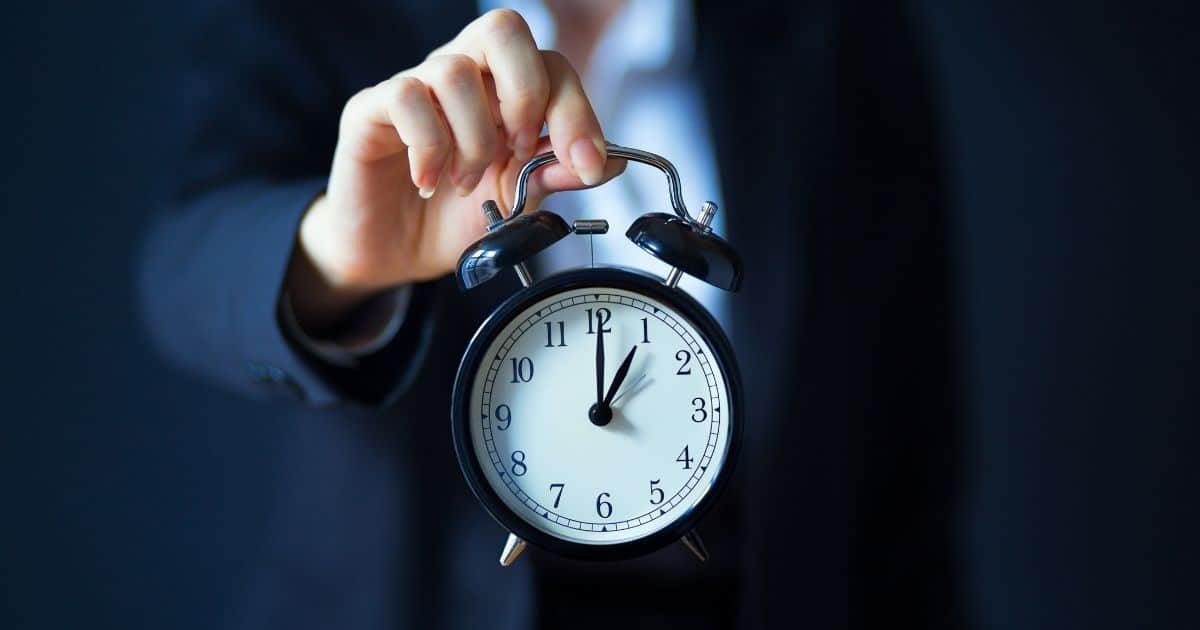 Een ouderwetse blauwzwarte wekker geeft aan dat het 1 uur is. Een dame houdt de wekker vast met haar duim en wijsvinger. Haar bovenlichaam is vervaagd zichtbaar op de achtergrond. Ze draagt een donkerblauw maatpak met licht hemd.