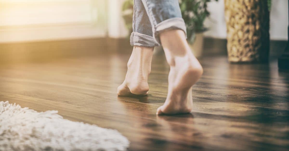 Blote voeten lopen over een warme houten vloer. Op de vloer ligt een wit hoogpolig tapijt. De voeten steken uit een jeansbroek met opgerolde pijpen.