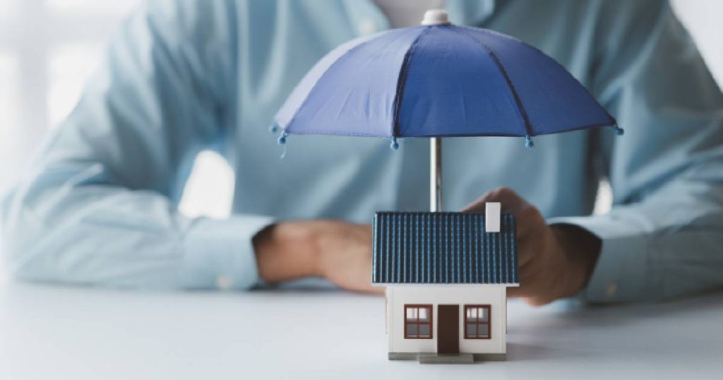 Un homme en chemise bleue tient un petit parapluie bleu au-dessus d'une petite maison blanche et bleue : un symbole pour comparer les polices d'assurance incendie pour protéger votre maison et vous-même contre les dommages et les coûts élevés.