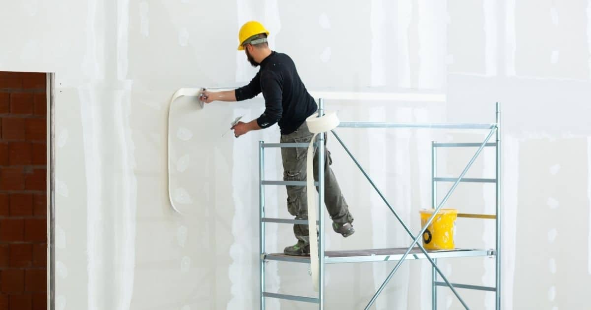 Een stukadoor staat op een steiger en bereidt de muren van een nieuwbouwwoning voor op pleister- of stucwerken. Naast hem op de steiger staat een gele emmer. De man draagt een gele veiligheidshelm, zwarte trui en grijsgroene werkbroek.