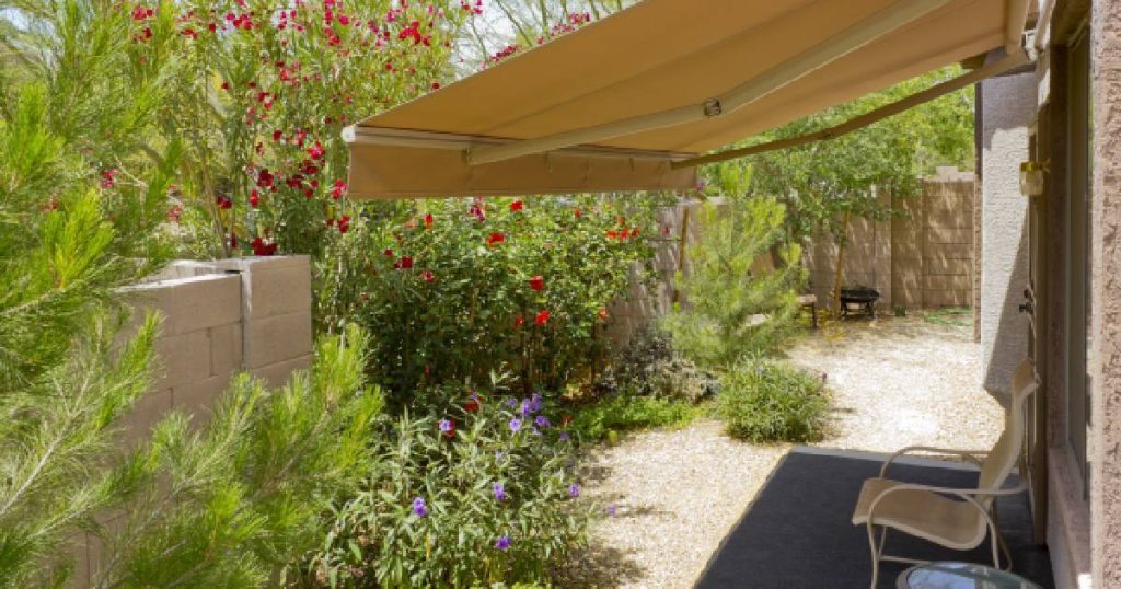 Een buitenzonwering voor een minimalistische veranda in een kleine tuin. De zonnewering bestaat uit een warmgeel zeil of doek op een manueel te bedienen frame. Onder de verandazonwering staat een tuinstoeltje. Voor de zonnewering staan verschillende grote struiken met bloemen.