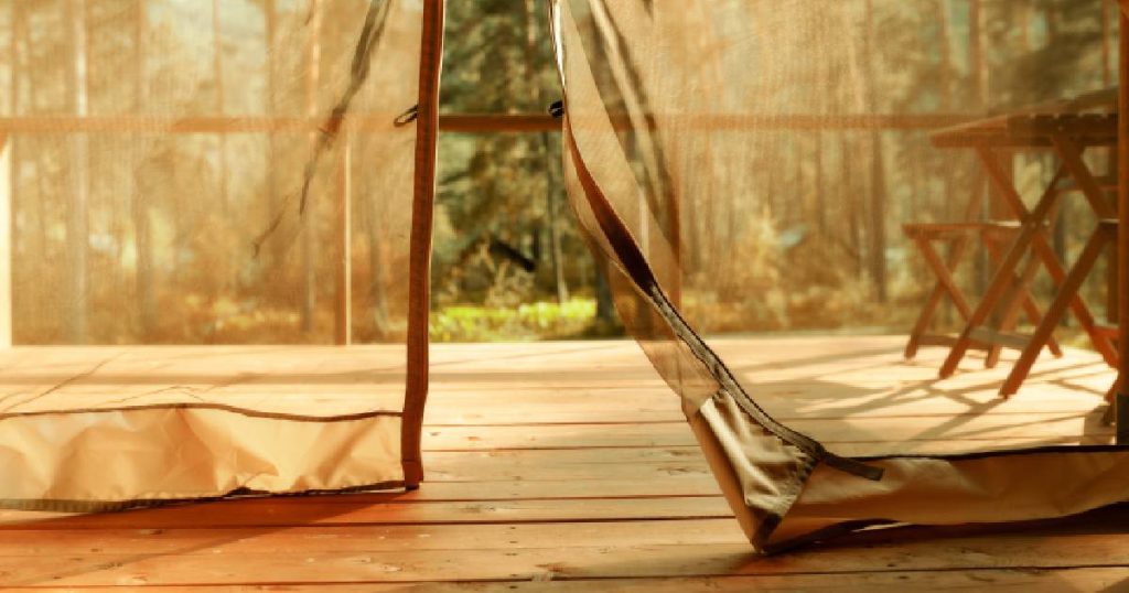 Een veranda met houten vloer, afgewerkt met een buitenzonwering van licht doorlatend zeil. Achter het zeil loopt de houten vloer door. Door het zeil zijn een houten klaptafel en klapstoel zichtbaar, in de verte op de achtergrond een bos.